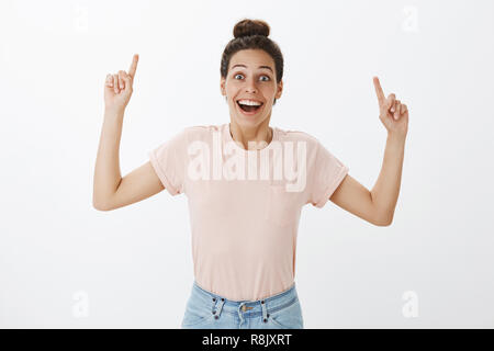 Waist-up shot of delighted enthusiastic surprised young happy girlfriend in casual t-shirt raising hands to point up smiling excited, joyful as reacting to awesome unexpected surprise over gray wall Stock Photo