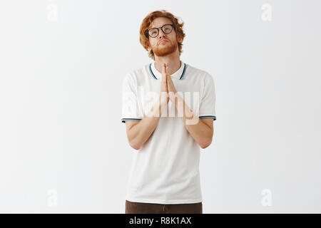 Studio shot of gloomy begging cute redhead guy with beard praying god help pass his exams holding hands in pray pursing lips and frowning while looking up with miserable expression Stock Photo