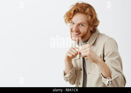 Redhead bearded man feeling thrilled before exciting boxing match standing in boxer pose with raised fists and broad thrilled smile as if fighting on ring over gray background in beige street jacket Stock Photo