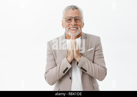 Cute and silly senior male in glasses and suit with white hair and beard holding hands in pray against chest clenching teeth saying please while begging for help or favour over grey wall Stock Photo