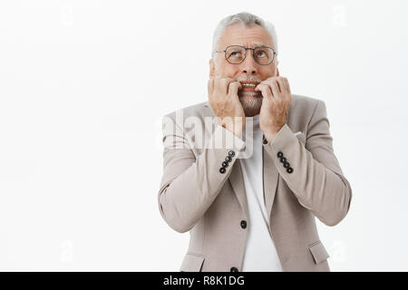 Portrait of concerned panicking and worried senior bearded man in glasses and suit biting fingers from nervousness and overreacting looking up scared standing intense over white background