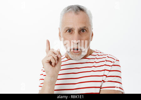 Excited creative and smart wise senior man with grey hair and beard in striped t-shirt raising index finger in eureka gesture folding lips, talking, adding suggestion over white background Stock Photo