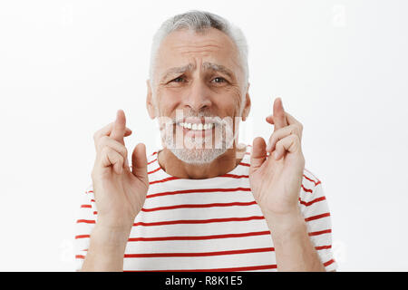 Intense worried senior retired grandfather with grey beard crossing fingers for good luck frowning clenching teeth praying making wish for wife getting cured standing nervous over gray wall Stock Photo