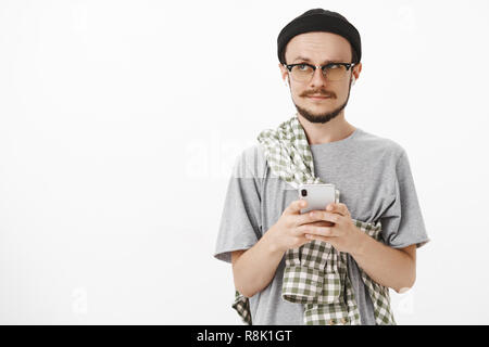 Waist-up shot of artistic questioned cute bearded male in black beanie and shirt crossed on chest holding smartphone raising eyebrow while gazing left with confused listening music in wireless earbuds Stock Photo