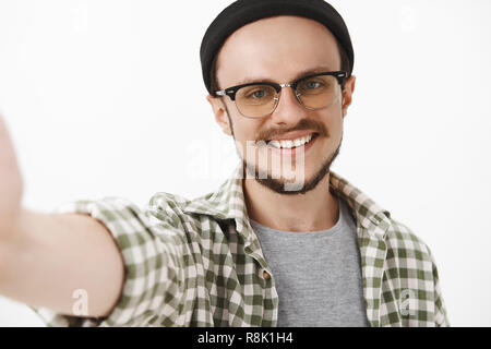 Close-up shot of friendly handsome neat guy in black trendy beanie and glasses with moustache smiling broadly pulling hand to hold camera while taking selfie on brand new smartphone Stock Photo
