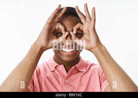 Close-up shot of funny and playful good-looking kind and happy african american guy showing okay gesture as binoculars on eyes smiling broadly looking on distance to find love of life over grey wall Stock Photo
