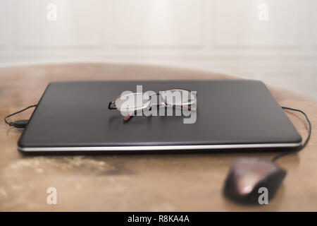 closed Laptop with eyeglasses on top and USB mouse on an old wooden table with white paneling in background Stock Photo
