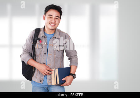 Photo image portrait of a cute young Asian male student standing, looking at camera and smiling while holding some books Stock Photo