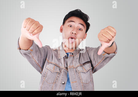 Photo image portrait of a cute young Asian man doing mocking gesture with tongue out and showing two thumbs down Stock Photo