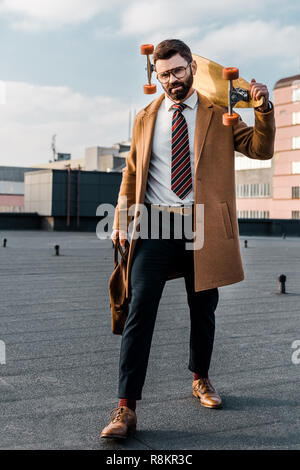 bearded businessman standing in formalwear and coat with penny board and bag in hands Stock Photo