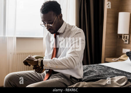 young african american businessman looking at wristwatch while sitting on bed in hotel room Stock Photo