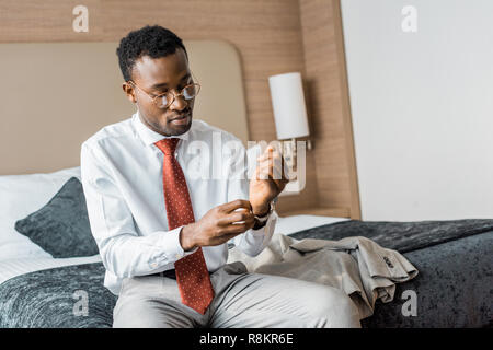 young african american businessman wearing wristwatch while sitting on bed in hotel room Stock Photo