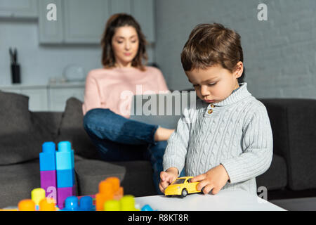 child playing with toy car while his mother using laptop behind at home Stock Photo