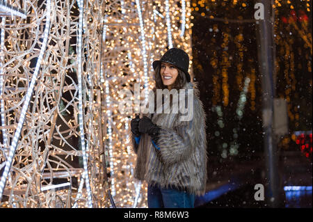 Pretty dark haired girl wearing a fur coat, blue jeans, blue top and a black hat, smiling, posing with snowflakes Christmas lights outdoor at night ti Stock Photo