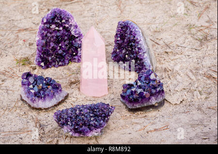 1 rose quartz gemstone surround by 5 amethyst crystals laying on the beach and in the grass Stock Photo