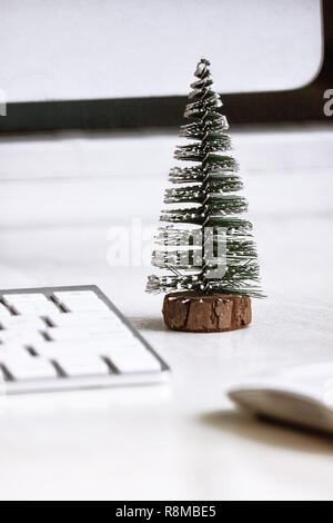 A small fir tree stands on a white desktop, near a computer monitor, keyboard and mouse. Concept, create a Christmas mood in the workplace. Stock Photo