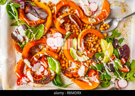 Baked pumpkin with chickpeas on a baking sheet with avocado, tahini and vegetables, top view. Stock Photo