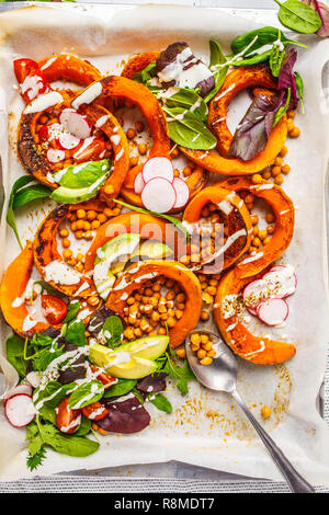 Baked pumpkin with chickpeas on a baking sheet with avocado, tahini and vegetables, top view. Stock Photo