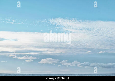 Cumulus and cirrocumulus clouds in the clear bright blue sky in the afternoon on the sea coast with a small strip of the sea below in the sunny day Stock Photo