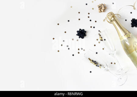 Happy New Year composition. Champagne wine bottle and glasses with golden confetti stars isolated on white table background. Party celebration concept. Flat lay, top view. Empty copy space. Stock Photo