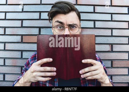 Funny face. Young hipster guy wearing eyeglasses with picked up one browe looking at camera and holding a book or note book on brick wall background. Stock Photo