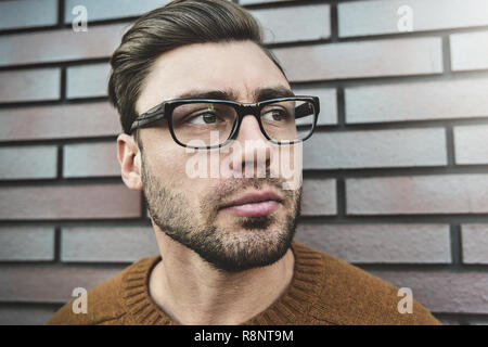 Portrait of serious manly stunning man in glasses and sweater looking at camera standing on street. Stock Photo