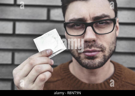 Contraception and healthy lifestyle concept. Male youngster with appealing look, holds condom. Bearded guy keeps contraceptive, advertises. Stock Photo