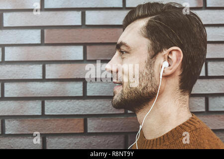 Photo of stylish handsome young man with bristle standing outdoors. Man wearing brown sweater. Smiling man listening to music on headphones and leanin Stock Photo
