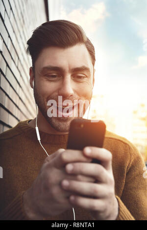 Smiling man listening to music on headphones and leaning against brick wall. Portrait of smiling man with headphones and cellphone standing by brick w Stock Photo