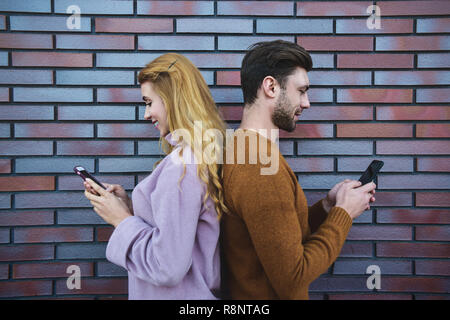 Side portrait of beautiful young couple is using smartphones and smiling, standing back to back against brown brick wall. Stock Photo