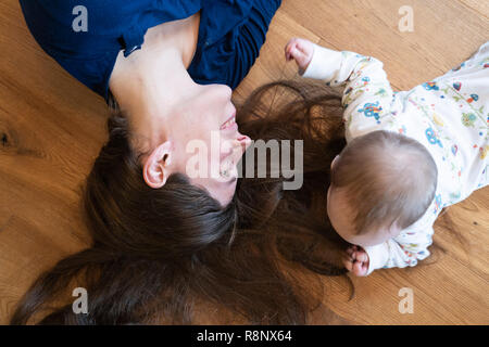 A baby girl lying on the floor and playing with her mother's hair with her mother smiling at her Stock Photo