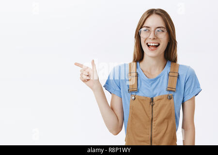 Waist-up shot of enthusiastic charming and charismatic happy girlfriend in brown overalls and transparent glasses pointing and looking left joyfully laughing having fun against gray background Stock Photo
