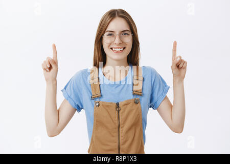 Waist-up shot of delighted carefree charming woman with friendly smile raising hands pointing up recommending place for cope space to entrepreneurs with positive charismatic attitude over white wall Stock Photo