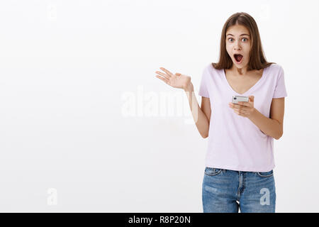 Amazed attractive sociable young female student with long brown hair holding smartphone waving raised palm and dropping jaw from awesome invitation to party received via interned over white wall Stock Photo