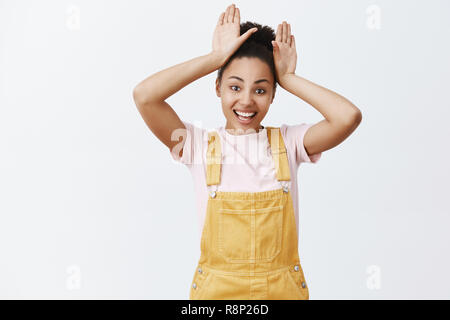 Studio shot of charming playful African American girl with combed hair in yellow overalls, holding palms on head and smiling broadly, mimicking bunny or puppy with long ears over grey wall Stock Photo