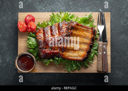 Closeup of pork ribs grilled with BBQ sauce and caramelized in honey on a bed of salad frisse. Tasty snack to beer on a wooden Board for filing on dark concrete background. Top view. Flat lay Stock Photo