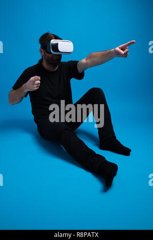 Male standing down and wearing the VR device pointing finger to the side. Wearing black clothes against blue background Stock Photo