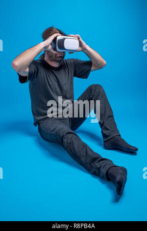 Portrait of a man standing down arranging his VR device. Wearing black clothes against blue background Stock Photo