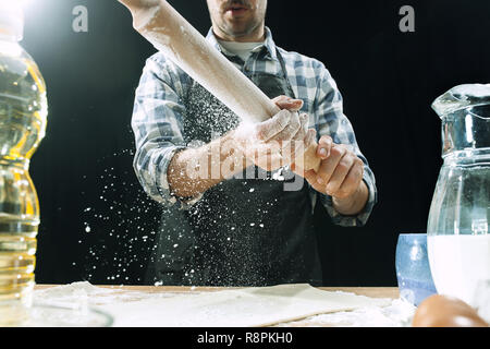 Professional male cook sprinkles dough with flour, preapares or bakes bread or pasta at kitchen table, has dirty uniform, isolated over black chalk background. Baking concept Stock Photo