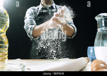 Professional male cook sprinkles dough with flour, preapares or bakes bread or pasta at kitchen table, has dirty uniform, isolated over black chalk background. Baking concept Stock Photo