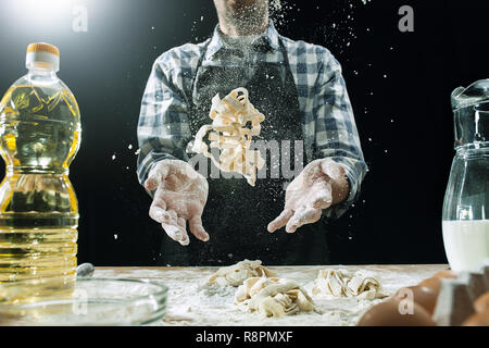 Professional male cook sprinkles dough with flour, preapares or bakes bread or pasta at kitchen table, has dirty uniform, isolated over black chalk background. Baking concept Stock Photo