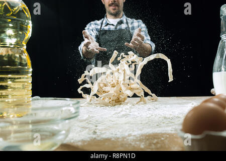 Professional male cook sprinkles dough with flour, preapares or bakes bread or pasta at kitchen table, has dirty uniform, isolated over black chalk background. Baking concept Stock Photo