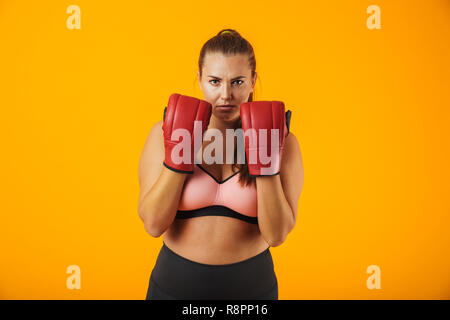 Portrait of sporty chubby woman in tracksuit wearing boxing gloves practicing isolated over yellow background Stock Photo