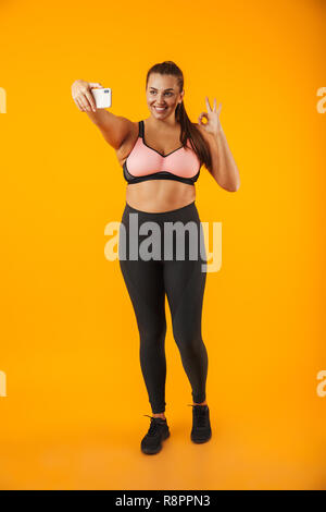 Full length portrait of cheerful chubby woman in sportive bra taking selfie photo on mobile phone isolated over yellow background
