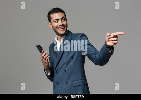 Image of joyful arabic businessman 30s in formal suit smiling while holding cell phone isolated over gray background Stock Photo