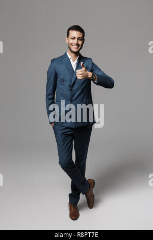 Full length image of cheerful arabic businessman 30s in formal suit gesturing at camera isolated over gray background Stock Photo