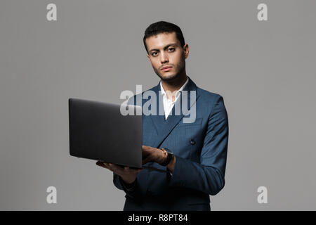 Portrait of a confident young businessman dressed in suit standing isolated over gray background, using laptop Stock Photo