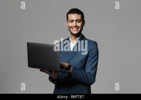 Portrait of a smiling young businessman dressed in suit standing isolated over gray background, using laptop Stock Photo