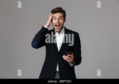 Image of delighted man 30s in business suit using black mobile phone isolated over gray background Stock Photo