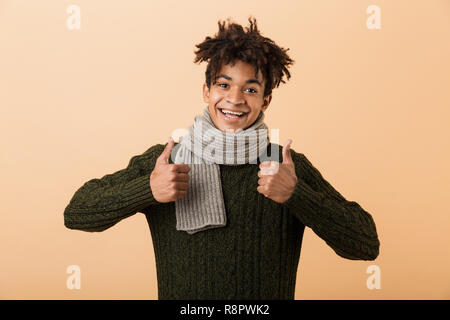 Portrait of cheerful african american guy wearing sweater and scarf showing thumbs up isolated over beige background Stock Photo